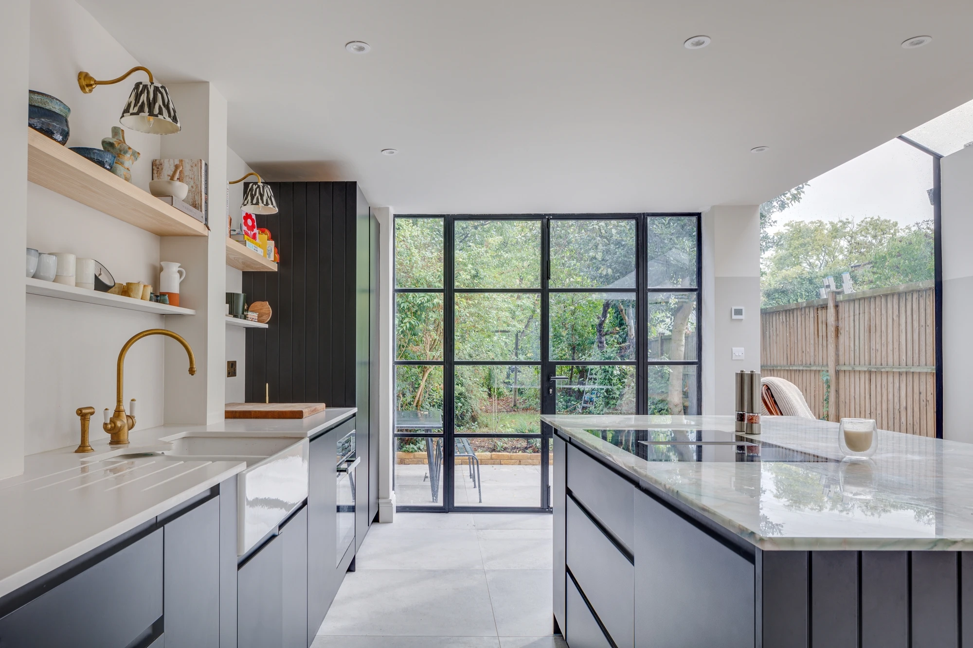 A modern kitchen with sleek gray cabinets, large windows, and a central island with a marble countertop. Open shelves display various kitchen items. The room is bright with natural light from the expansive glass doors opening to a garden