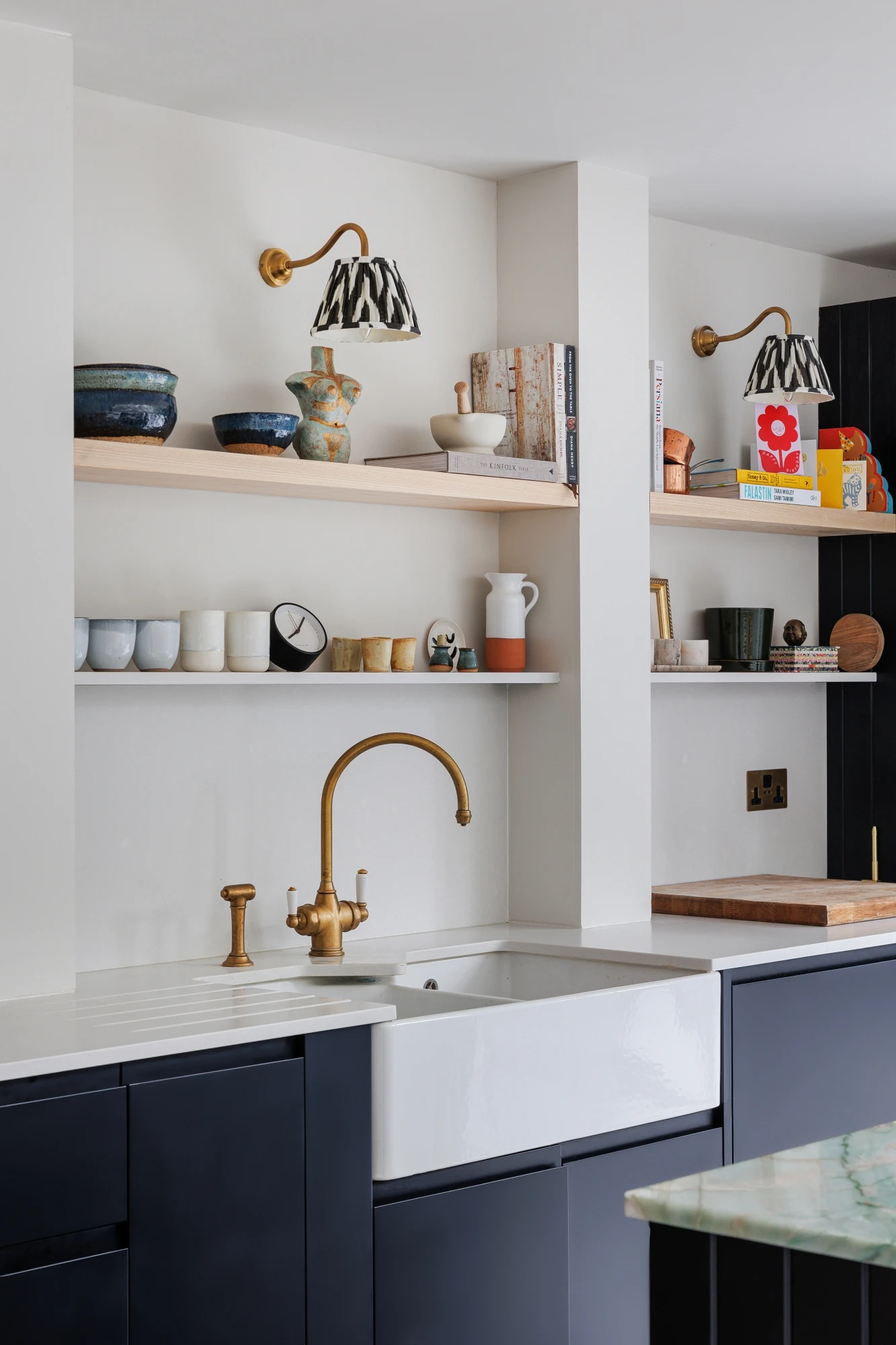 Modern kitchen with navy cabinets, a farmhouse sink with a brass faucet, and white countertops. Two shelves display various ceramic dishes, pots, and books. Two gold wall sconces with black and white patterned shades are mounted above the shelves.