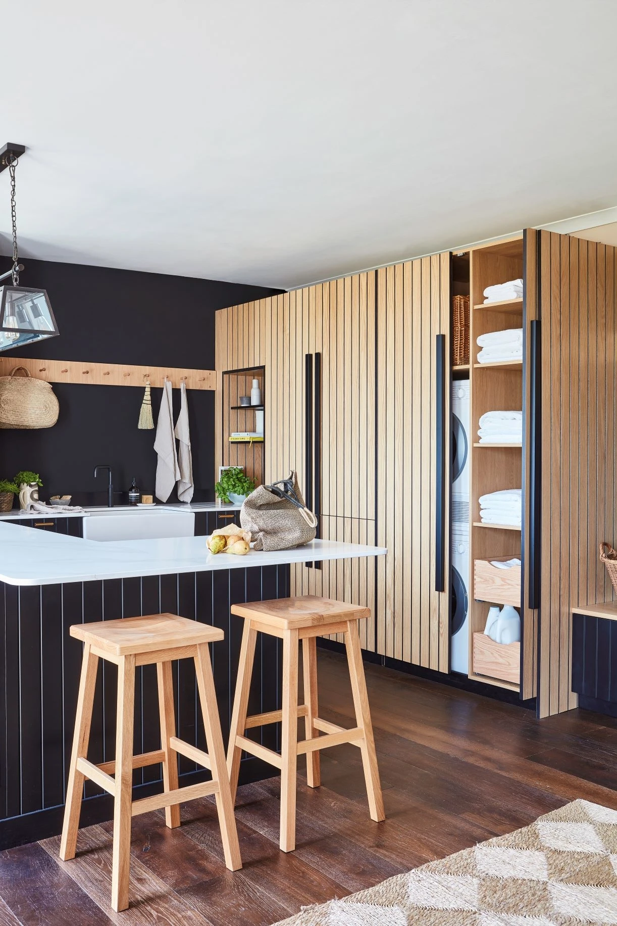 A modern kitchen with wooden vertical paneling on cabinets, a kitchen island with a white countertop, and two wooden stools. Open shelving displays folded towels. The flooring is dark wood, and the walls are painted black.