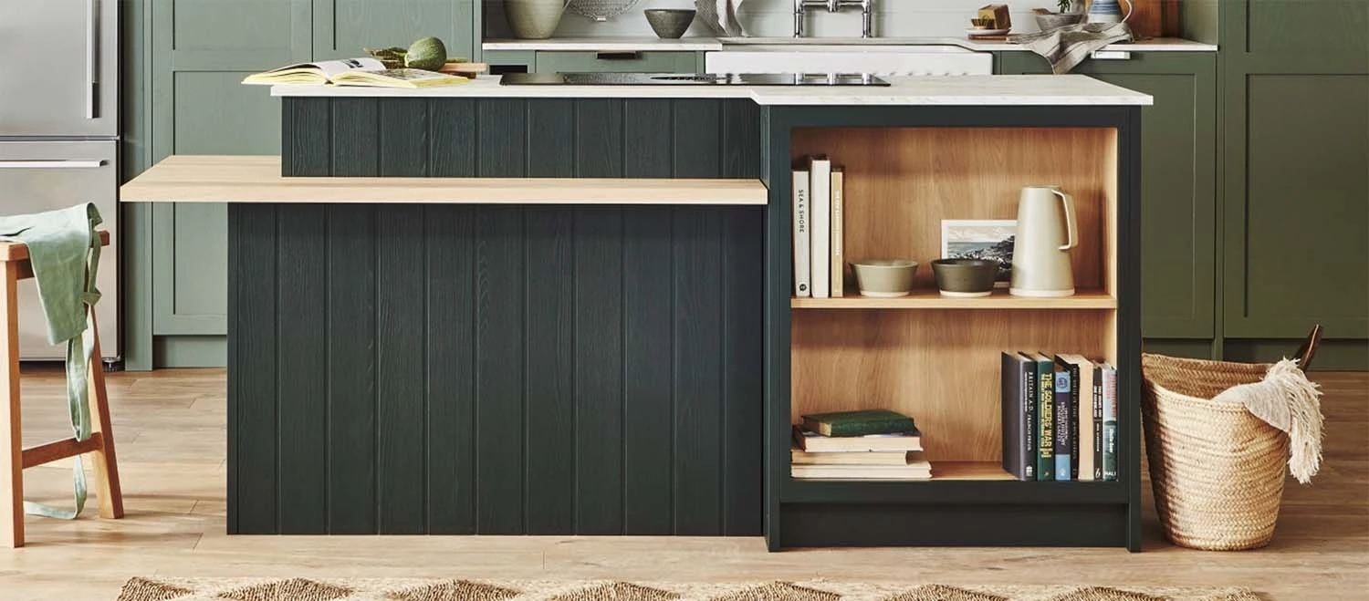 Modern kitchen with green cabinets, a black kitchen island with a wooden shelf displaying books and dishes, and a basket on the floor. The countertop has a sink and various kitchen items. Light wood flooring completes the cozy look