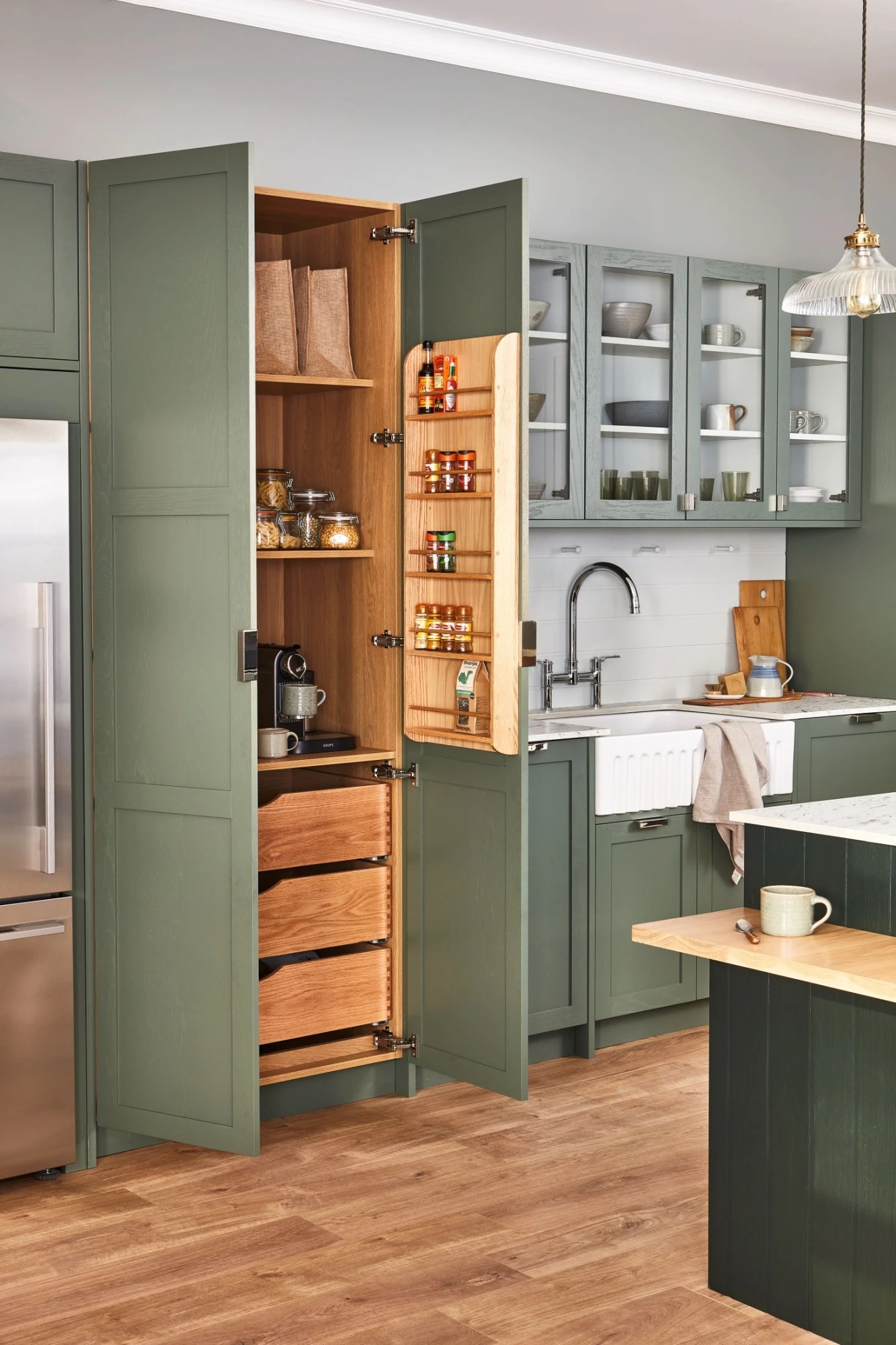 A modern kitchen with green cabinets and light wood floor. The open pantry reveals shelves with various items, including jars of spices, bowls, and paper bags. A stainless steel refrigerator is on the left, and a white farmhouse sink with a silver faucet is on the right.