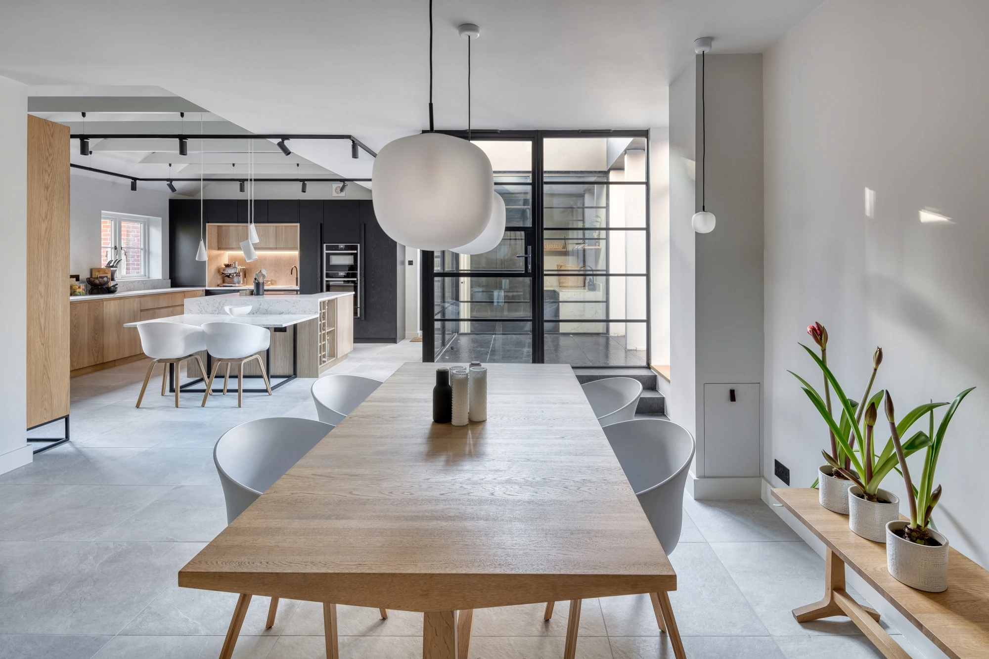 A modern kitchen and dining area featuring light wood furniture and minimalist decor. The dining table is accompanied by white chairs and large pendant lights hanging above. The kitchen has an island in the center, with a mix of open and glass-fronted shelving units.