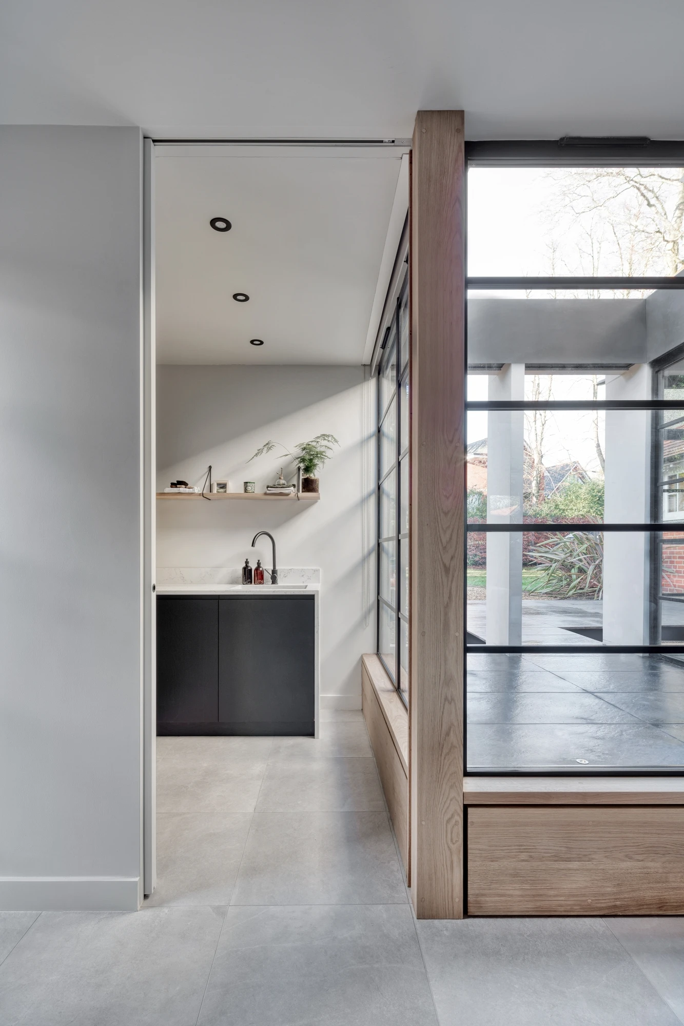 Modern interior showing a sleek kitchen with minimalist black and white design, including a black sink and white countertops. Adjacent to the kitchen is a bright, glass-walled living area with wooden trim, leading out to a garden with green foliage and a brick wall