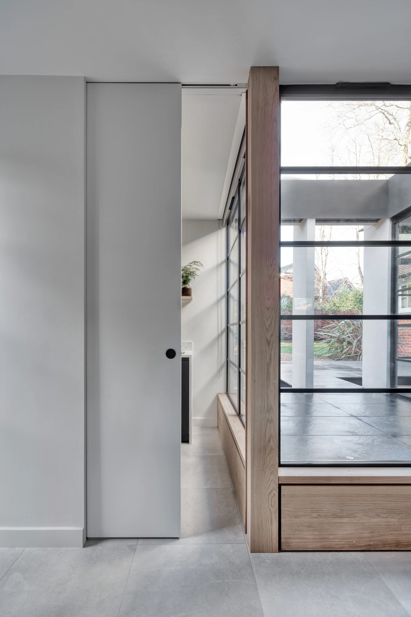 Modern interior showing a sleek kitchen with minimalist black and white design, including a black sink and white countertops. Adjacent to the kitchen is a bright, glass-walled living area with wooden trim, leading out to a garden with green foliage and a brick wall