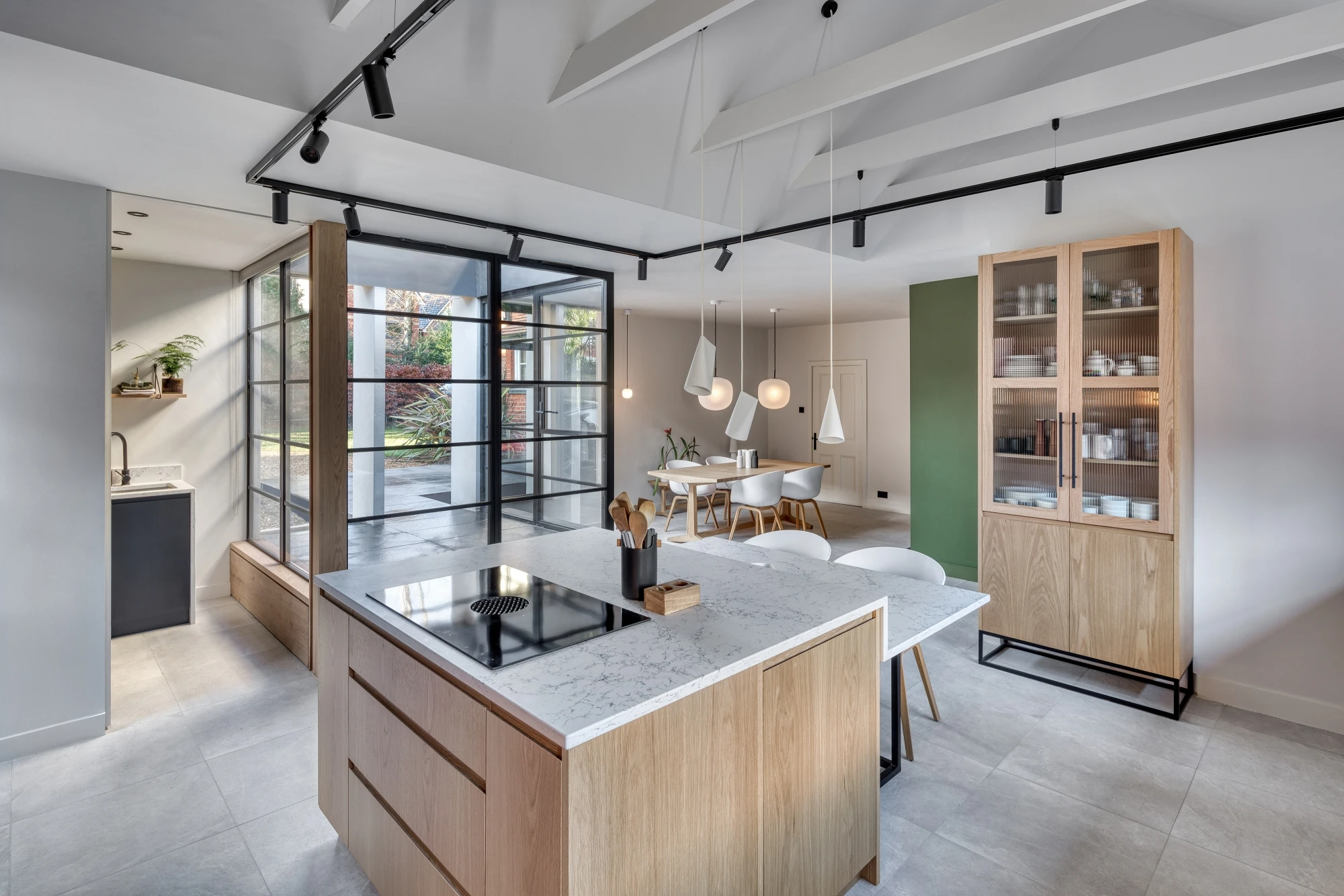 Modern open-concept kitchen and dining space with light wood cabinetry and white countertops. The kitchen has a kitchen island with an induction cooktop and ceiling-mounted track lighting. Large windows and glass doors offer ample natural light and a view of greenery