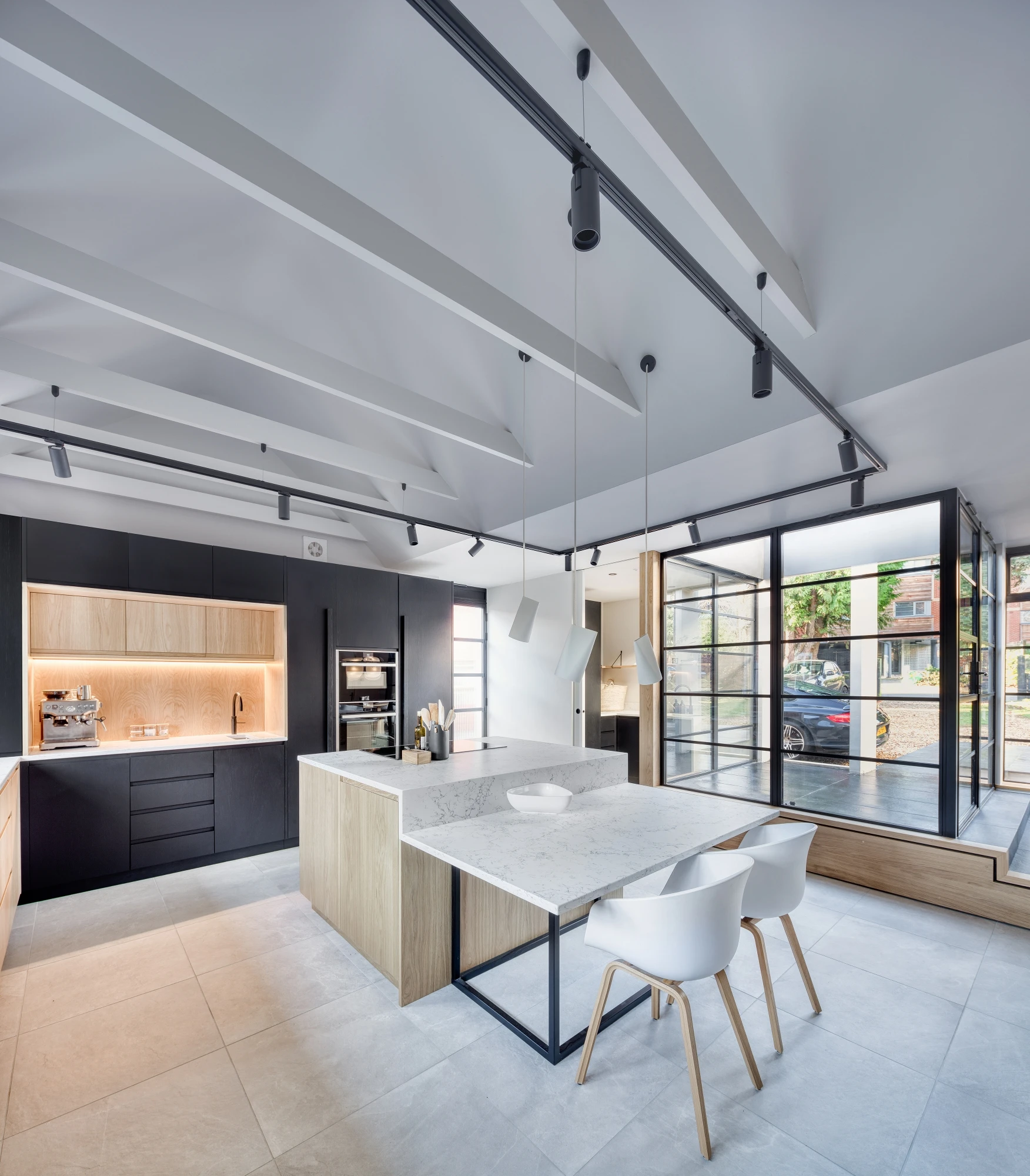 A modern kitchen with a minimalist design. It features a central island with white countertops and two white chairs. The surrounding cabinetry is a mix of black and light wood. Large windows allow natural light, and exposed beams and track lights decorate the ceiling