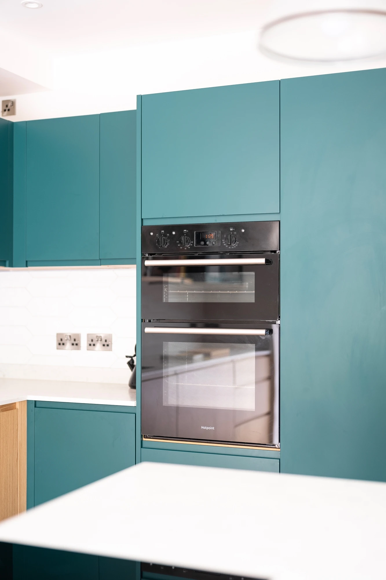 A modern kitchen with turquoise cabinets featuring a built-in black oven and microwave. The countertop is white with a kettle placed on it. The backsplash has a white honeycomb tile design, and there are power outlets on the wall.