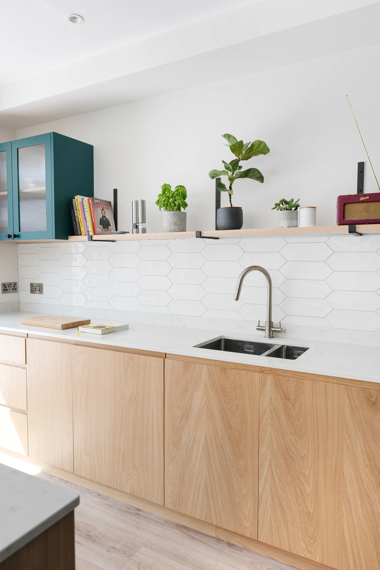 A modern kitchen with light wood cabinetry, white hexagonal tile backsplash, and a white countertop. Stainless steel sink with a tall faucet. Green and white potted plants, books, and decorative items are arranged on open shelves above the counter