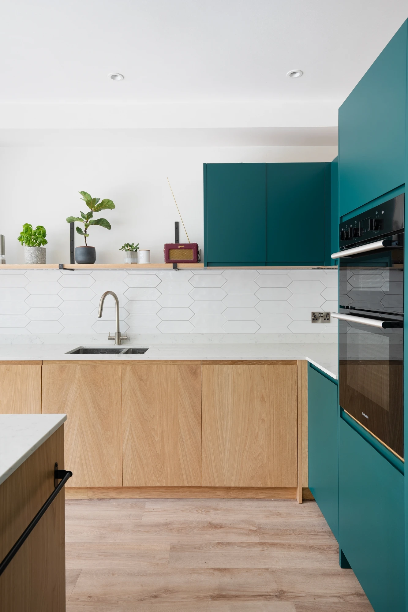 A modern kitchen with a mix of teal and wooden cabinetry. The backsplash features white hexagonal tiles. There's a stainless steel oven and microwave on the right and a sink on the wooden countertop to the left. Potted plants decorate the shelf above the sink