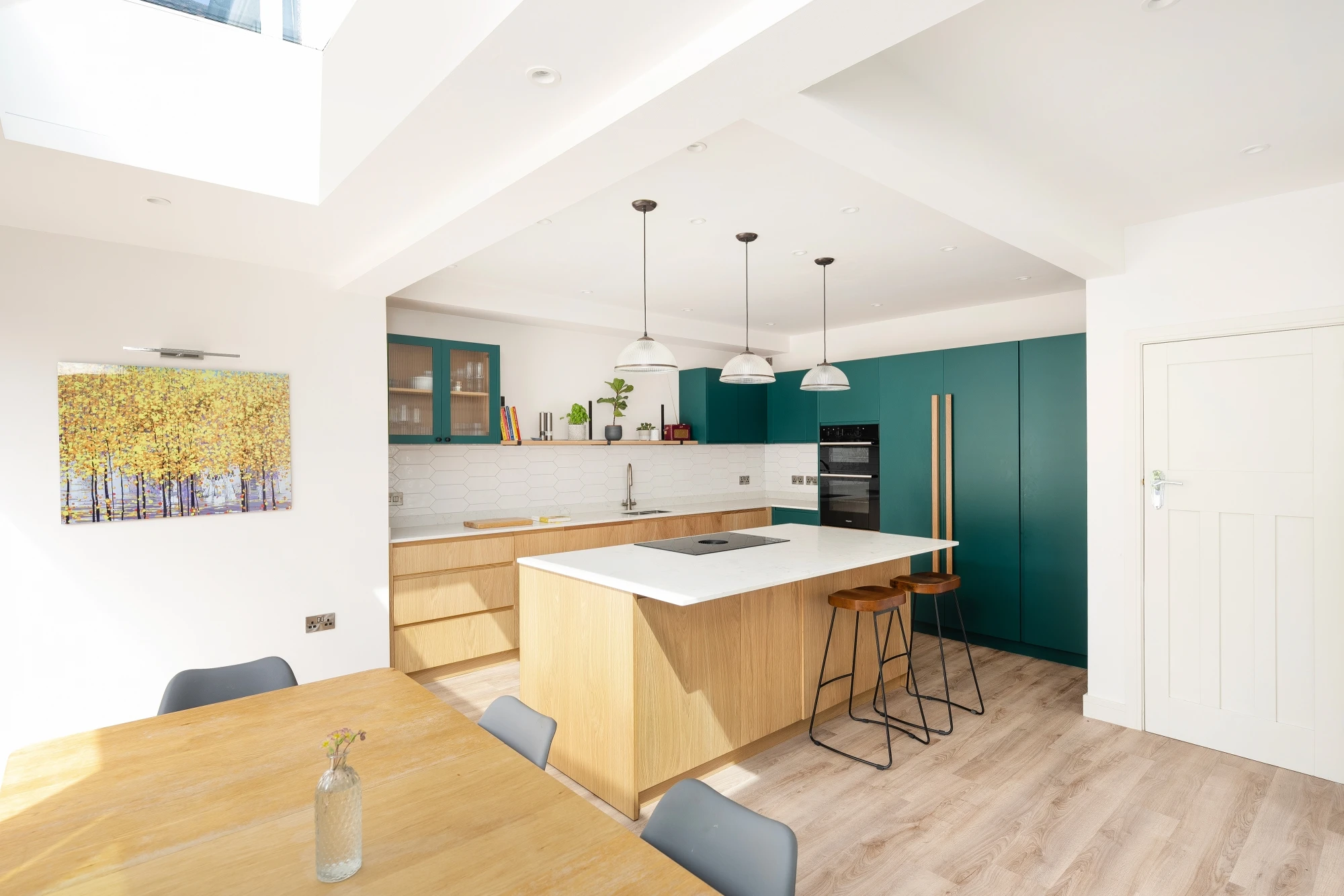 Contemporary kitchen with light wood cabinetry, white countertops, and teal accents. Pendant lights hang above a central island with seating. The open space features a dining table with chairs, hardwood flooring, and a framed autumn landscape painting on the wall