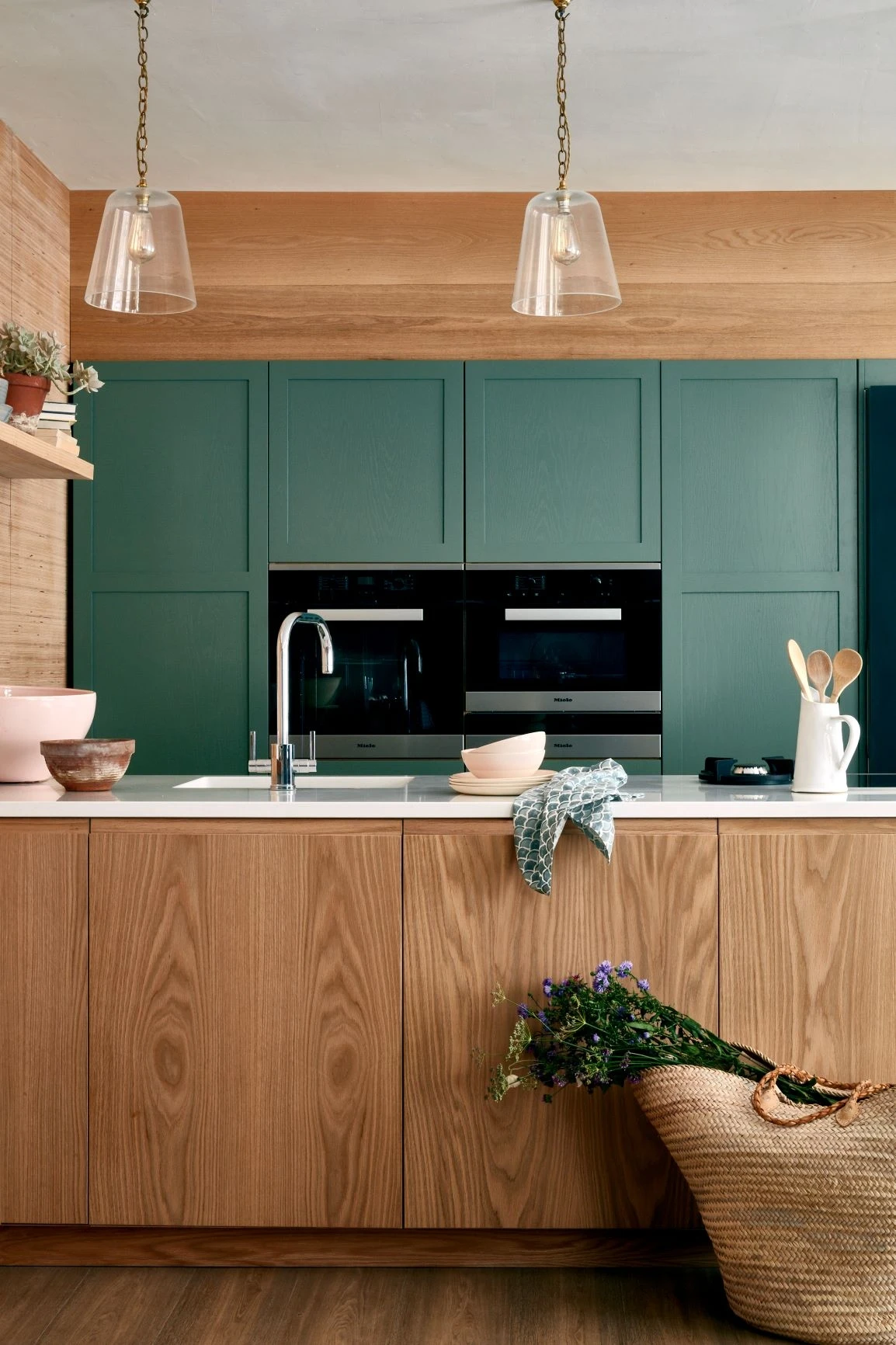 Modern kitchen with green cabinets and wood accents. The countertop features various kitchen items, such as bowls and utensils. Two pendant lights hang from the ceiling. A wicker basket with flowers is placed on the floor, leaning against the cabinets.