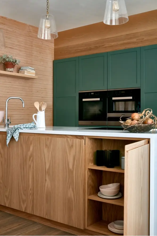 A modern kitchen with wooden cabinets and shelves. A white countertop features a faucet, a pitcher with utensils, and a basket of onions. Green cabinetry with built-in double ovens is in the background. Open shelves hold plates and cups, and pendant lights hang overhead.