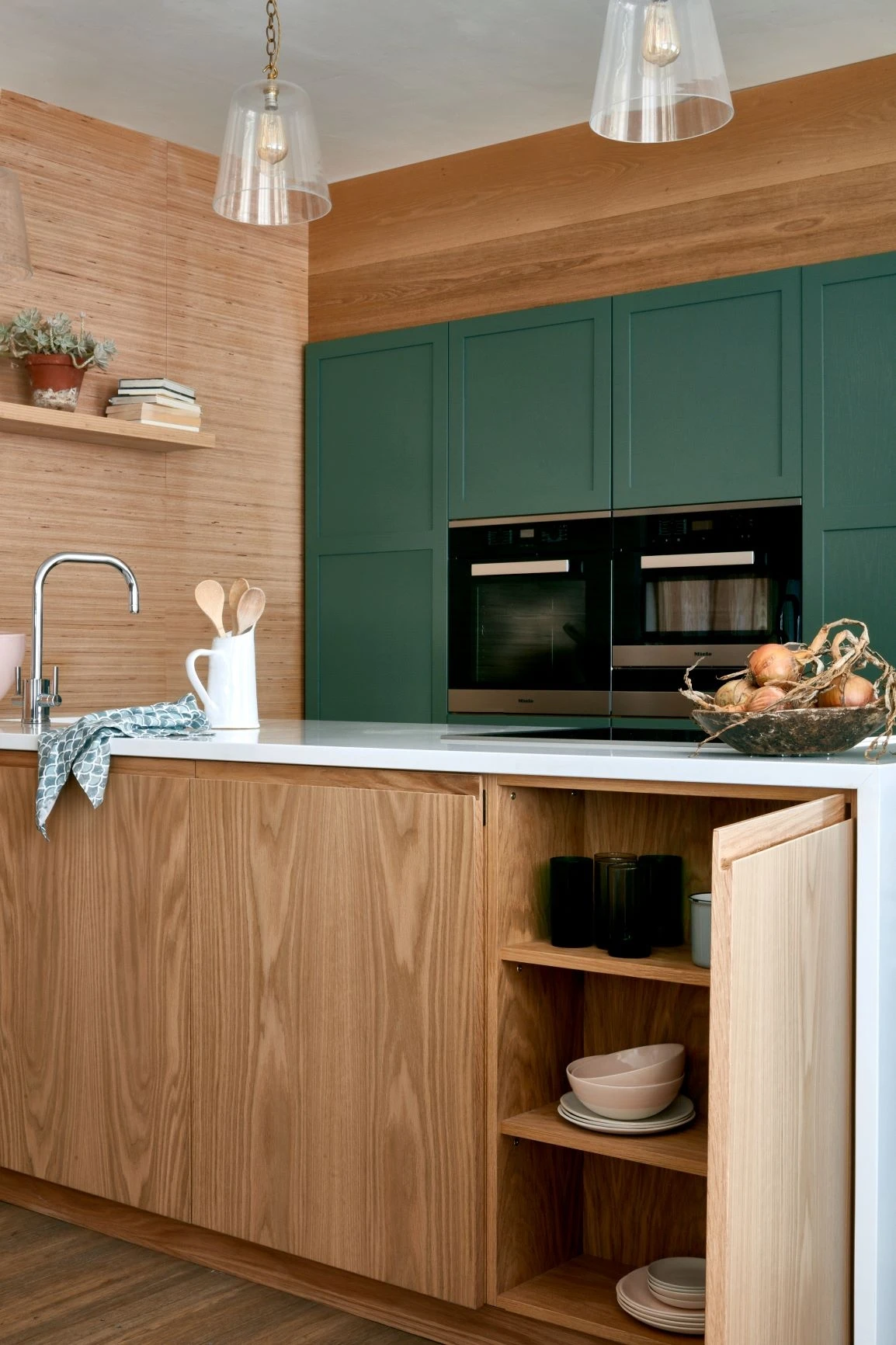 A modern kitchen with wooden cabinets and shelves. A white countertop features a faucet, a pitcher with utensils, and a basket of onions. Green cabinetry with built-in double ovens is in the background. Open shelves hold plates and cups, and pendant lights hang overhead.