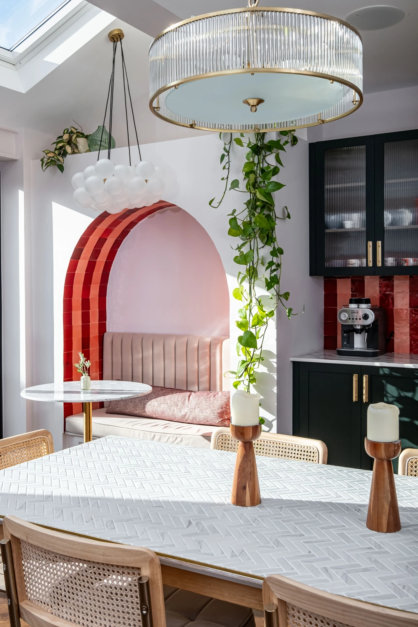 A bright, modern kitchen with a skylight. The room features a white dining table with candles, cane-back chairs, a cozy built-in bench with pink cushions under an archway, red tile backsplash, dark green cabinets, and hanging plants. Two elegant light fixtures hang from the ceiling.