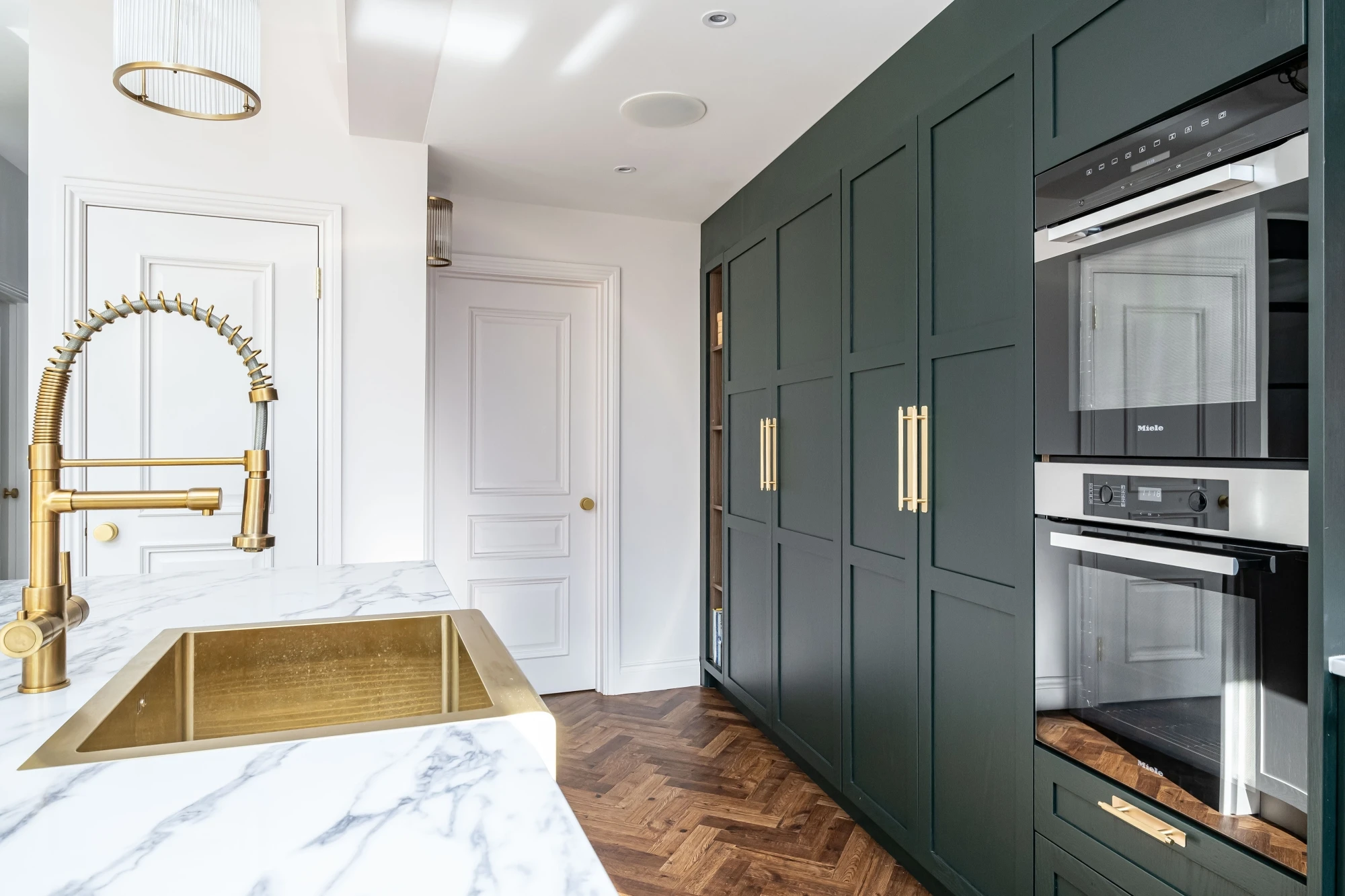 A modern kitchen featuring a marble countertop with a gold sink and faucet. Dark green cabinets with gold handles are on the right, housing built-in stainless steel appliances. The floor is patterned wood, and the background includes white doors and walls.