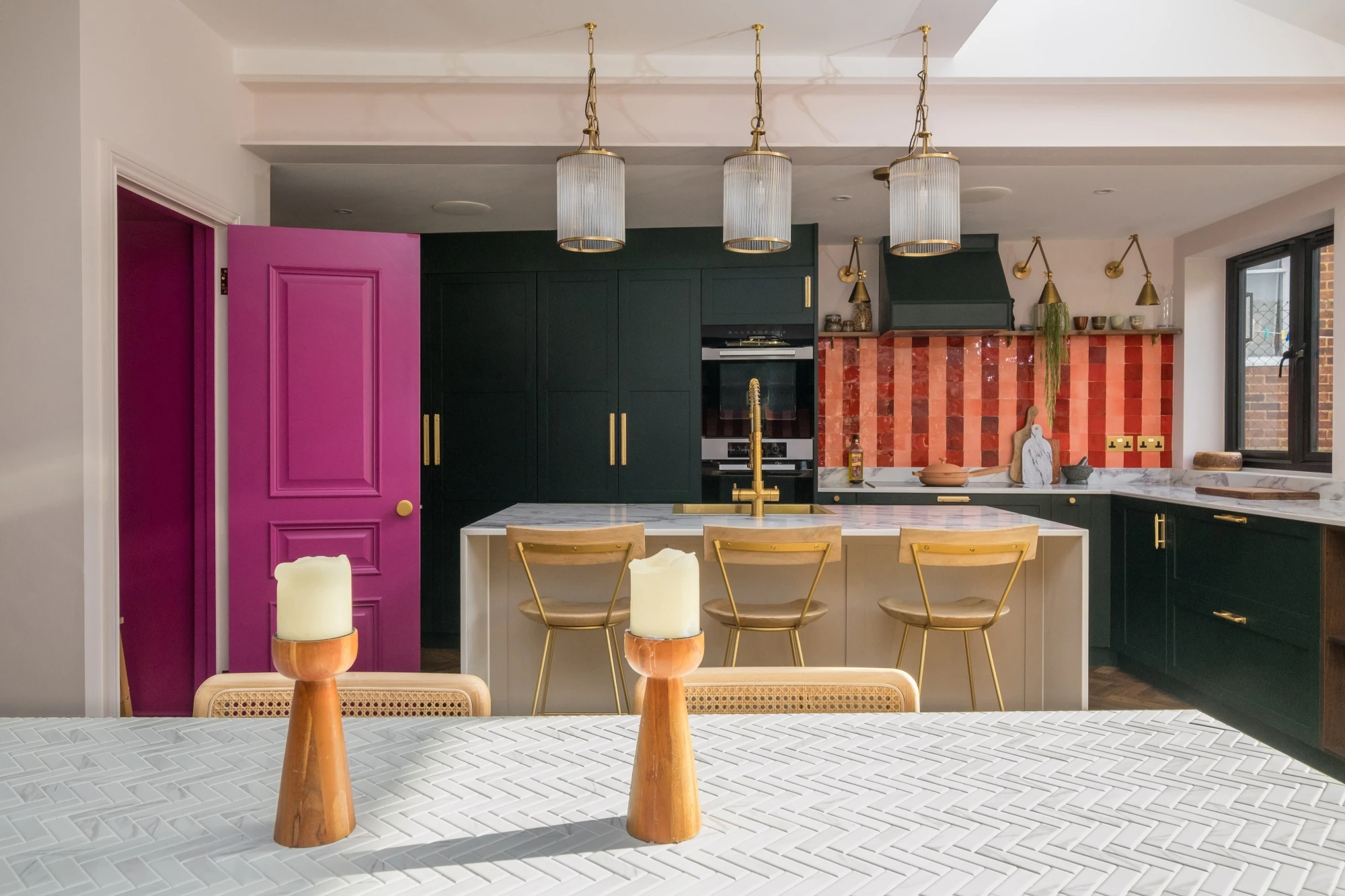 A modern kitchen with dark green cabinets, a central island with gold bar stools, and a colorful red and orange backsplash. Three glass pendant lights hang overhead, and two candles on wooden holders sit on a patterned white tabletop. A vibrant magenta door is open on the left.