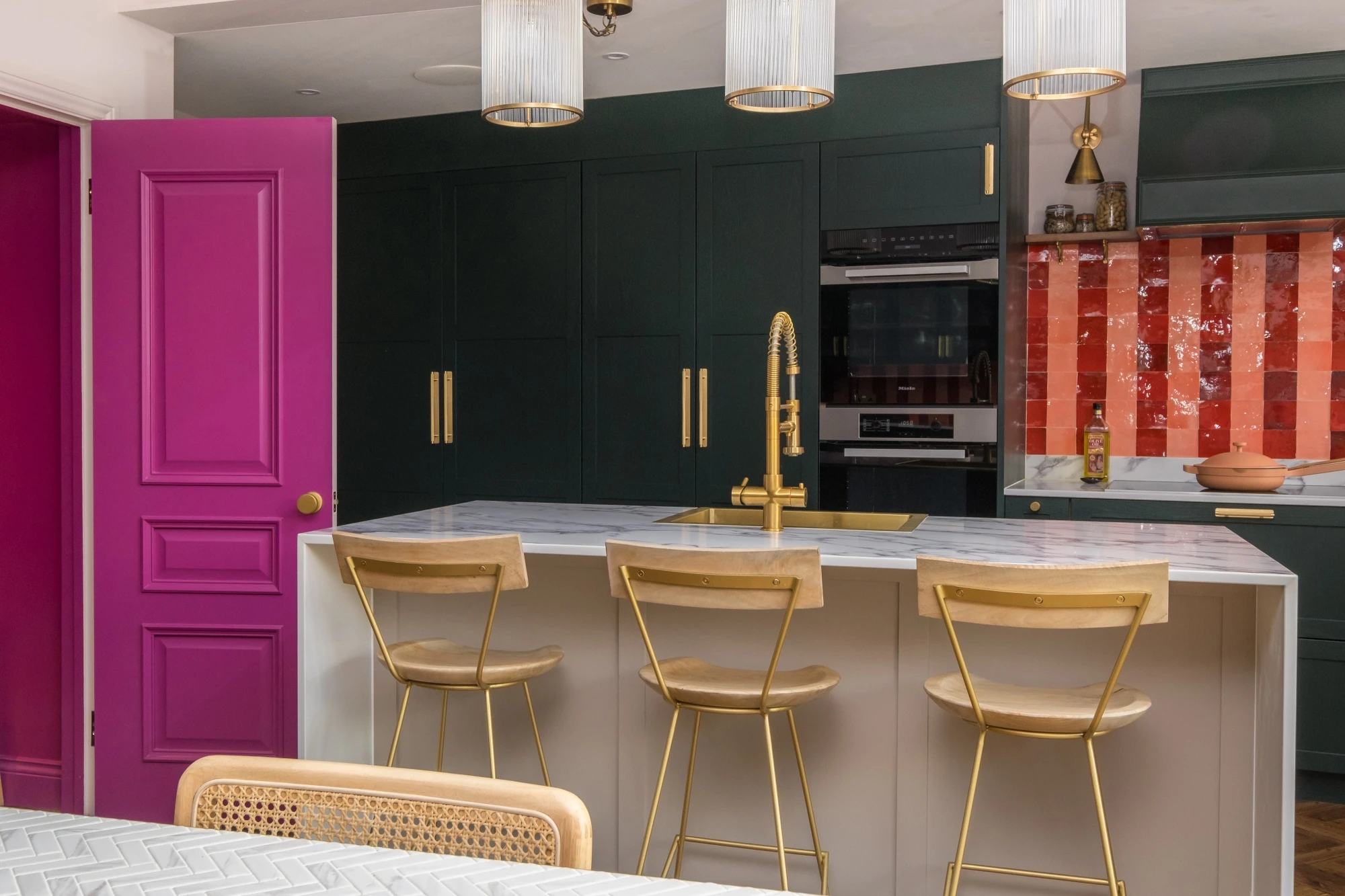 A modern kitchen features a central island with a white marble countertop, three gold metal chairs, a gold faucet, and dark green cabinets. A vibrant magenta door stands to the left, and a backsplash with orange tiles adds a pop of color behind the stove