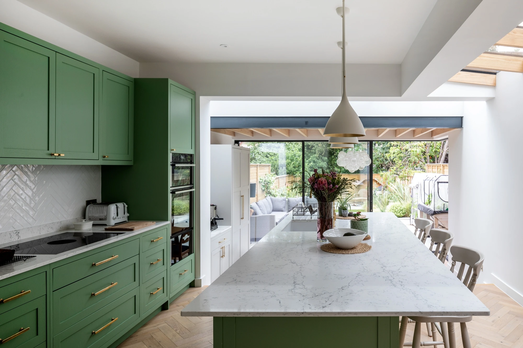 Modern kitchen with green cabinets, white marble countertops, and a large island. Features a pendant light, herringbone backsplash, and glass doors leading to an outdoor space.