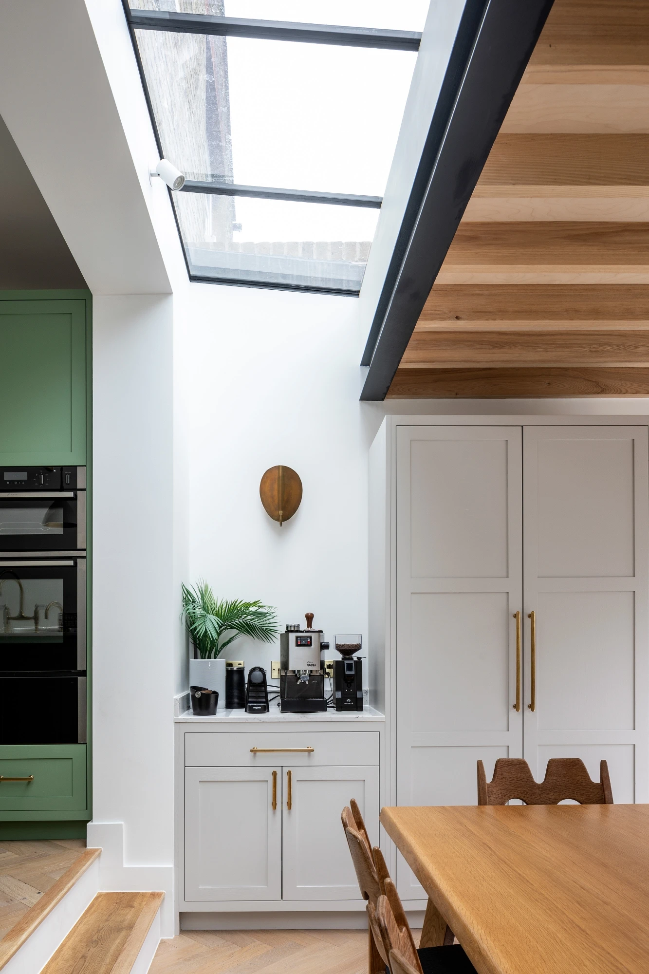 Coffee station with white cabinetry, gold handles, and a skylight above. Features a coffee machine, plant decor, and part of a dining table in the foreground.