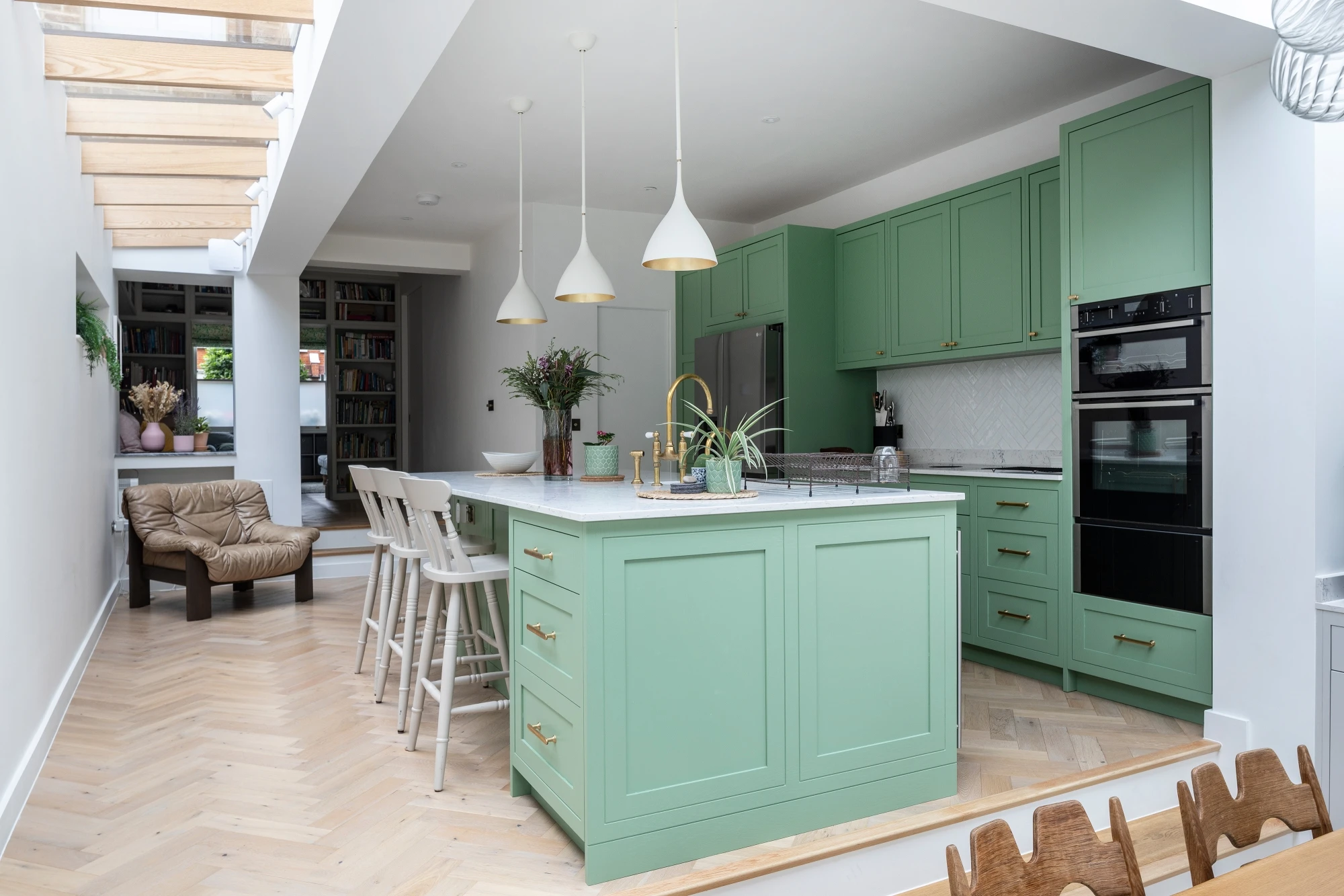 Bright kitchen with green cabinets, white island, and gold accents. Features pendant lights, a herringbone wood floor, and a cozy seating area with a tan chair.