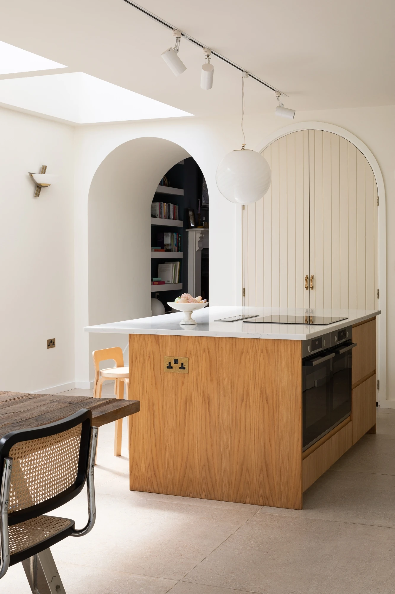 A modern kitchen featuring a wooden island with a white countertop. An induction cooktop and built-in oven are integrated. A large white spherical pendant light hangs above. An archway in the background leads to a cozy nook with bookshelves.