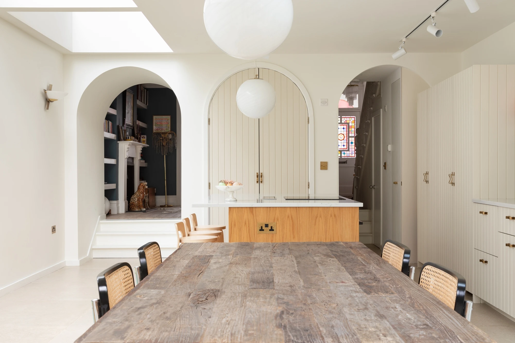 A modern dining area featuring a rustic wooden table with wicker-back chairs. The space includes a kitchen island with a white countertop, arched doorways, and a mix of contemporary and classic décor elements. A large floating white light fixture hangs from the ceiling