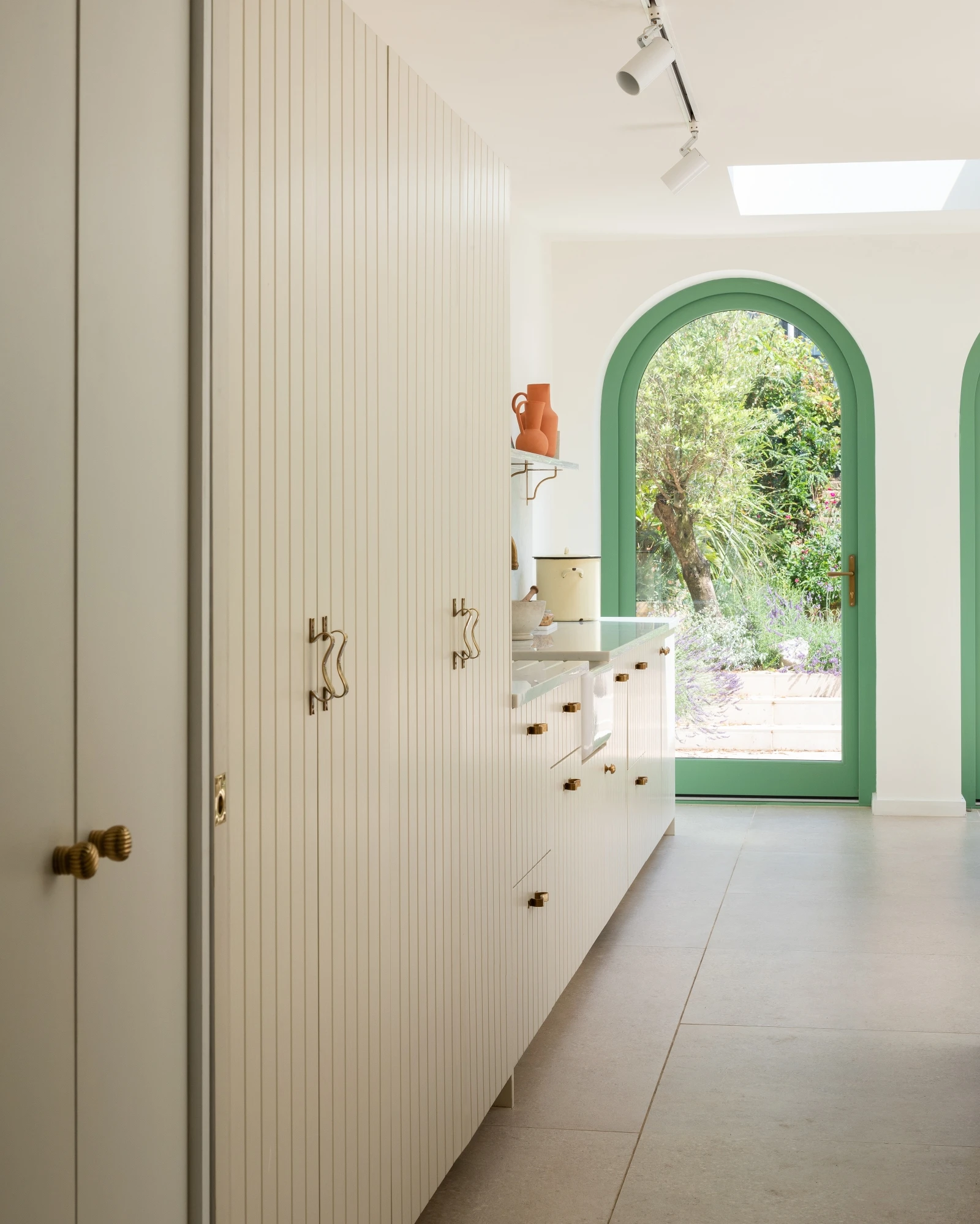 A modern kitchen with light-colored vertical wood panel cabinets and brass hardware. Two green arched doorways open to a lush garden. A skylight brightens the space, and the minimalistic design includes clean lines and neutral tones.