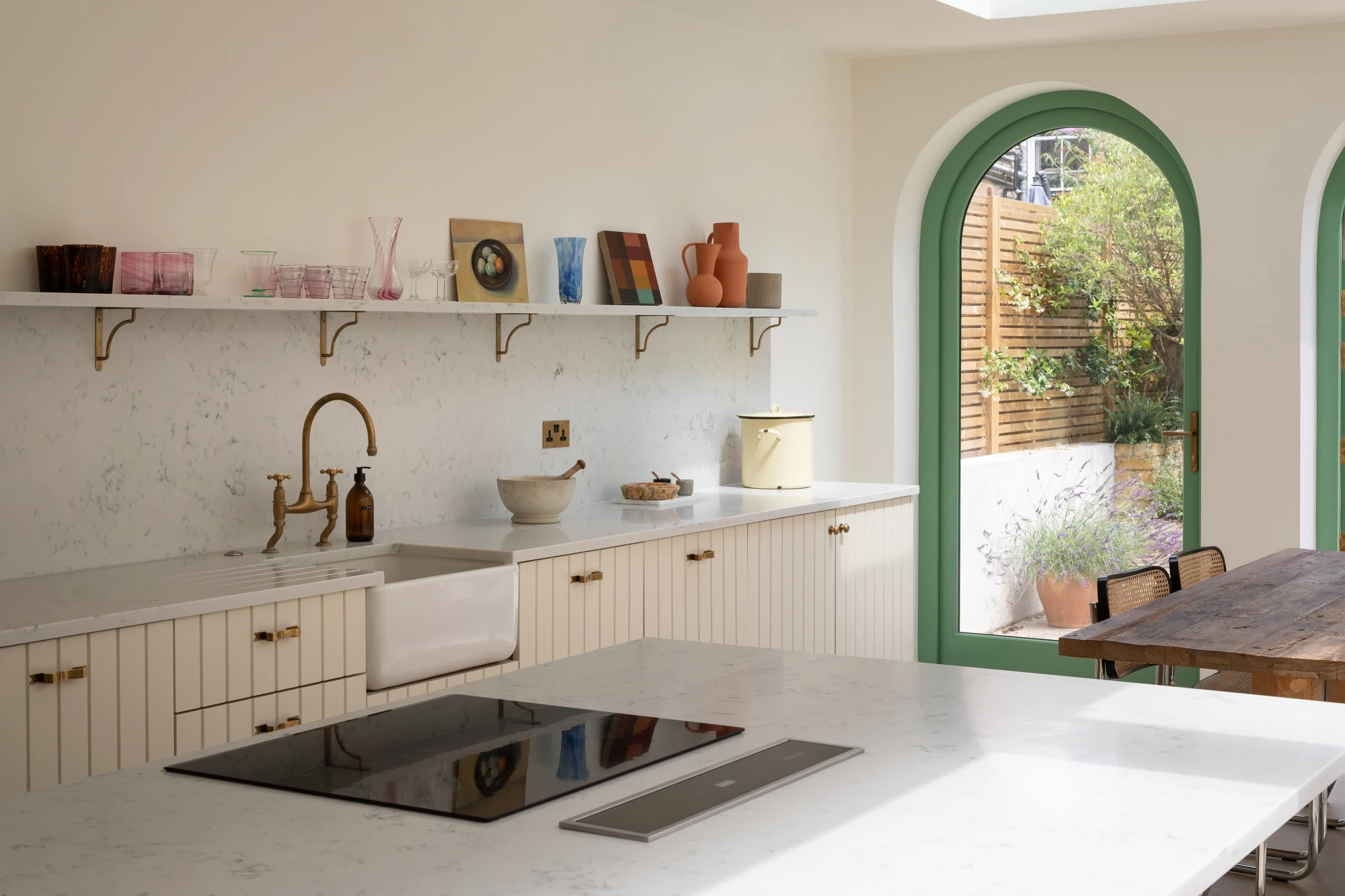 A modern kitchen with a marble countertop island featuring a black stovetop. The countertop against the wall includes a farmhouse sink and brass faucet, with shelves holding decorative items above. A large arched window and sliding door provide natural light