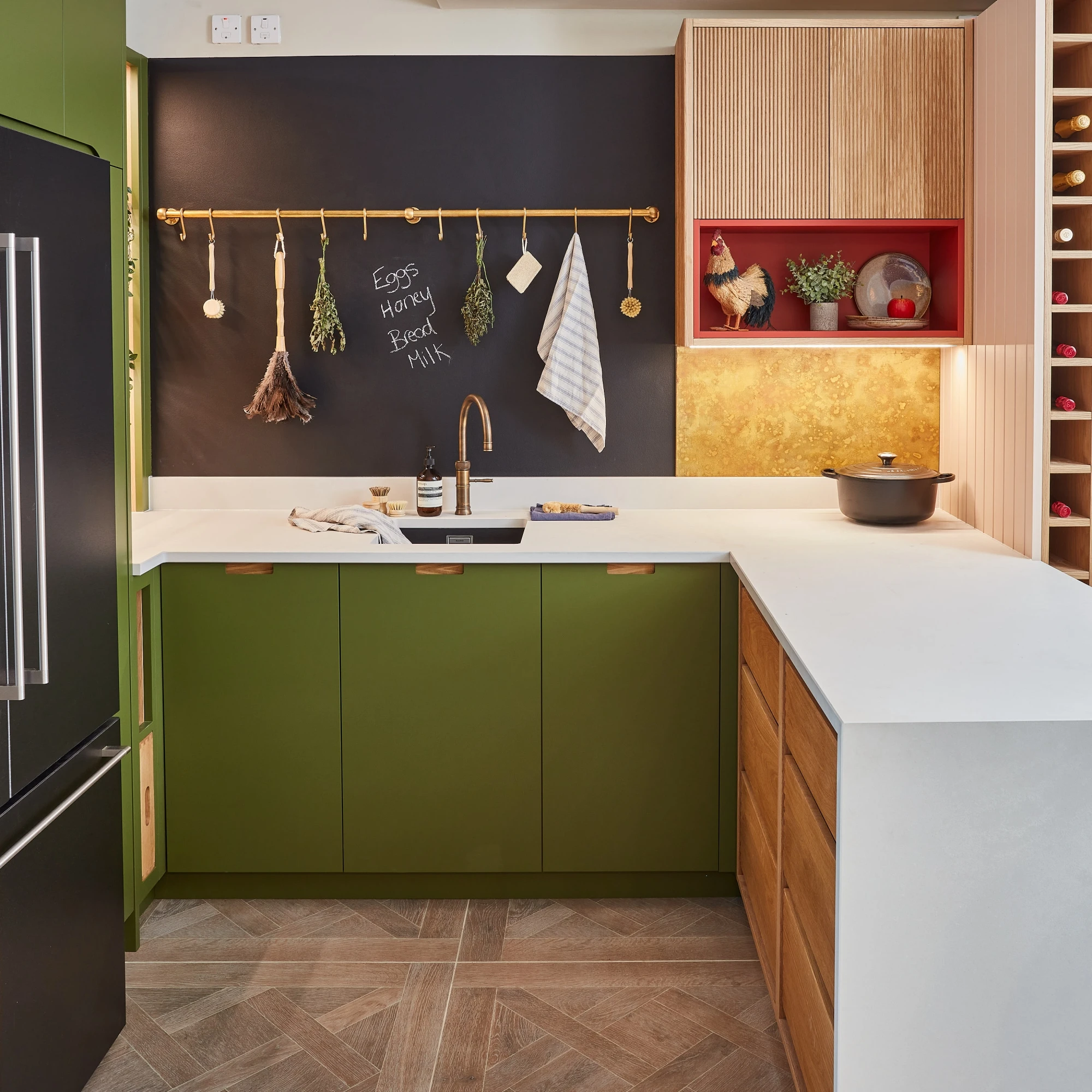 A modern kitchen with olive green and wood cabinetry, a white and gray marble countertop, and a light brown textured rug. Two pendant lights hang over an island. Beige brick and marble backsplash add contrast. Two framed pictures are on the wall.