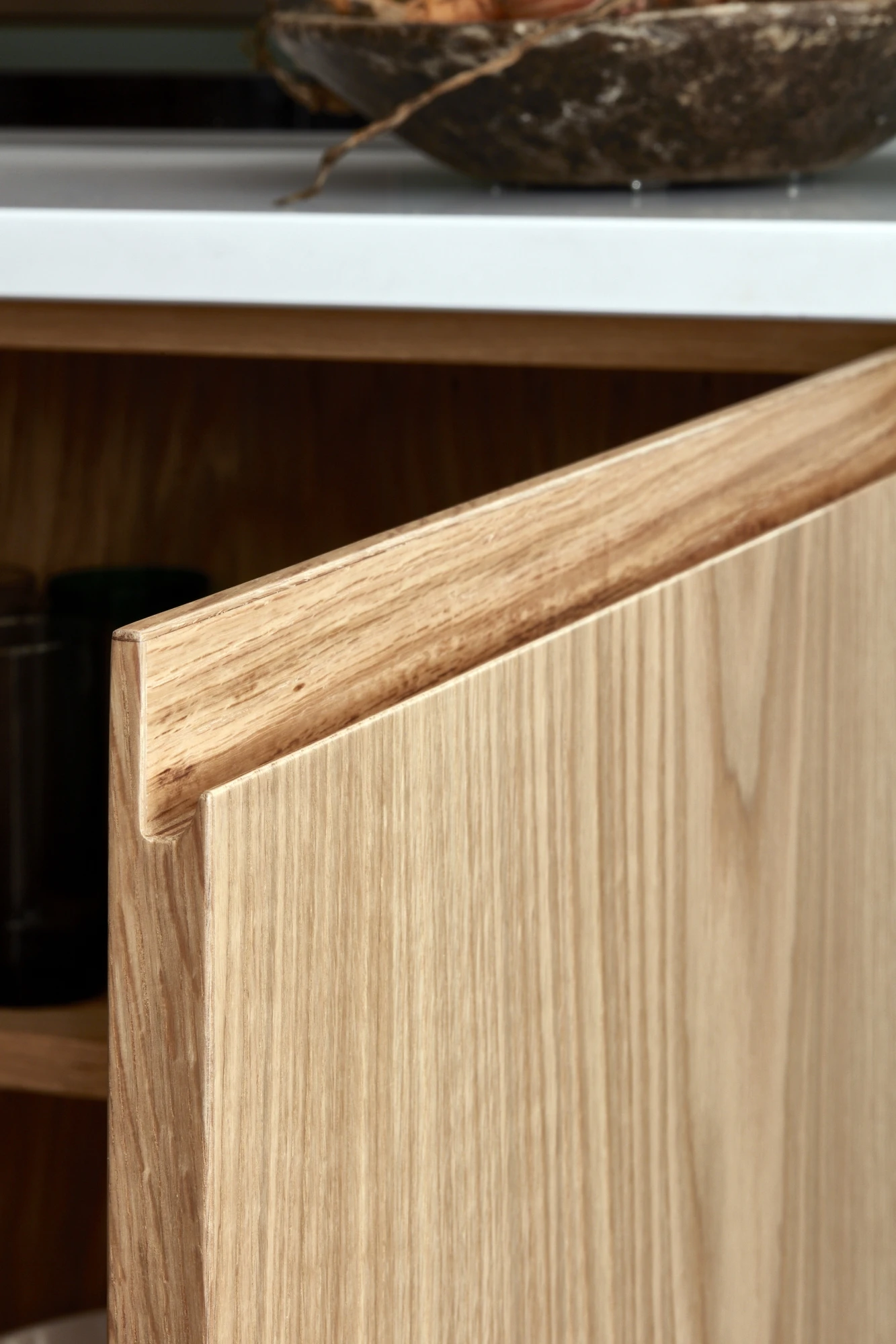 Close-up of a partially open wooden cabinet door with a simple, modern design. The background shows a white countertop with a shallow bowl partially in view on top. The cabinet door features a natural wood grain finish.