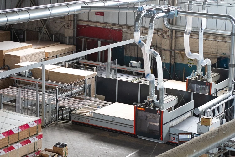 The image shows an industrial factory interior with large CNC machines processing wood panels. Stacks of cut wood are organized nearby, and air ducts hang from the ceiling. The space looks spacious and organized for manufacturing.