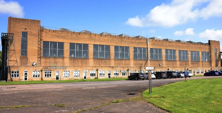 A long, rectangular industrial building with large windows and a flat roof. The foreground shows a paved area with parked cars and a grassy lawn. The sky is blue with a few clouds.