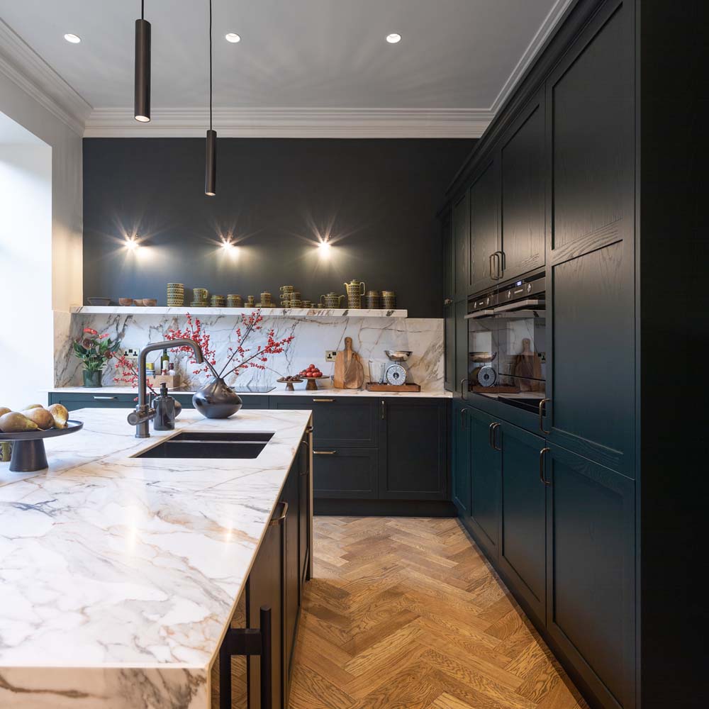 An image of a large kitchen with a penisula island. Marble worktops and splashbacks, with contemporary shaker cabinetry in a custom paint colour