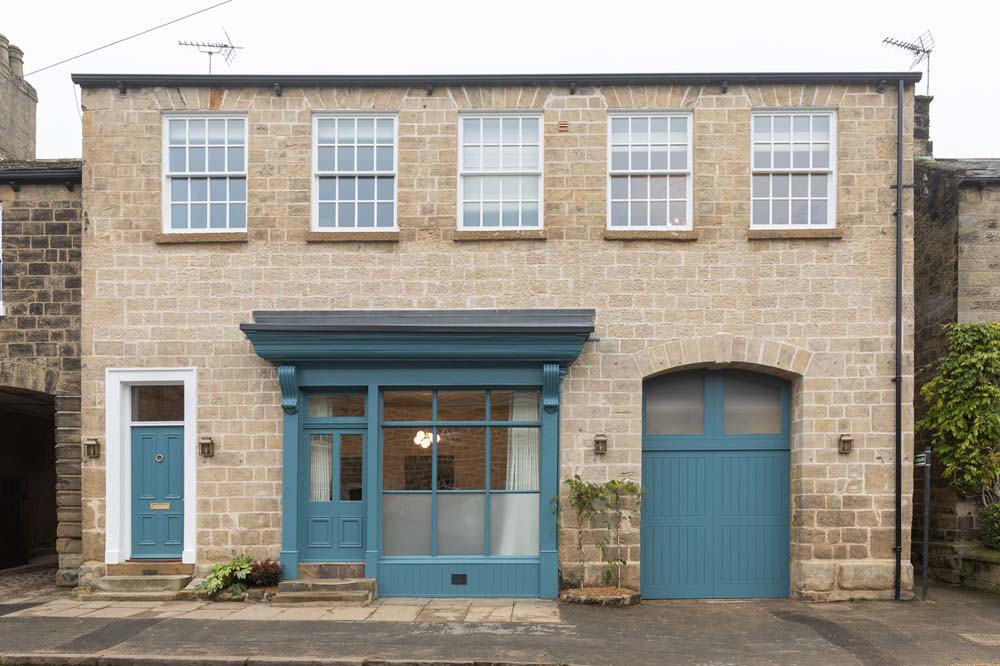 A photograph of the outside of Dawn's house. There is brickwork, with 5 upstairs windows, a door to the left, the old shopfront still visible in the middle and a gateway to the right. The door, shop front and gateway are painted in a blue colour, and the window frames and door trims are painted white.